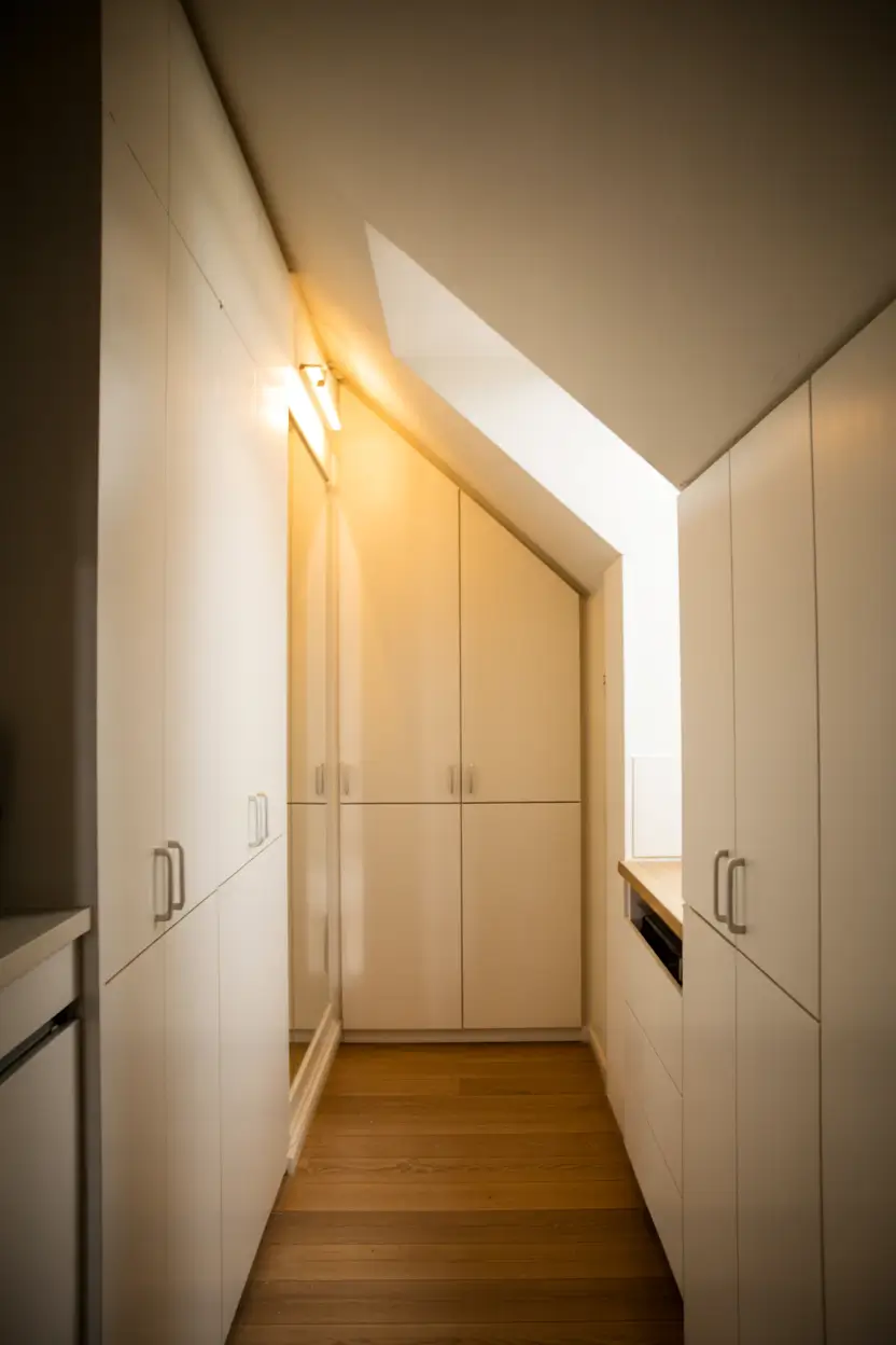A photograph of a compact Scandinavian hallway with sloping ceilings. The hallway features custom-built white cabinetry that fits seamlessly into the eaves, optimizing storage space. The cabinetry has sleek, minimalist handles, and the walls are painted a soft gray, creating a clean and airy atmosphere. Warm, natural light filters through a small window, casting gentle shadows and highlighting the hallway's functional yet stylish design.